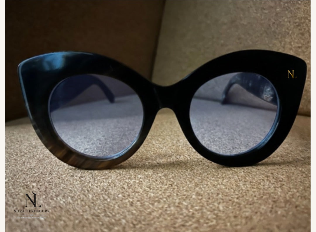 Black cat-eye frame in cow horn with dark tinted lenses and a small gold 'NL' logo on the upper right frame, resting on a textured beige surface.
